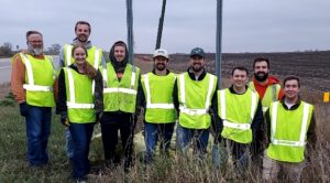 Nine Christianson employees in neon pose in front of an 'adopt a highway' sign