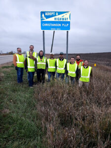 Nine Christianson employees in neon pose in front of an 'adopt a highway' sign