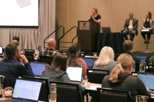 A woman gives a presentation to diverse group of professionals engaged in discussion while working on laptops in a modern conference room setting.