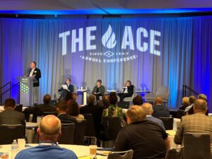 A man gives a presentation at the Ace Annual Conference on stage in front of a blue curtain.
