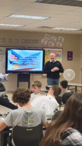 A man stands at the front of a classroom, presenting to an engaged group of students seated at desks.