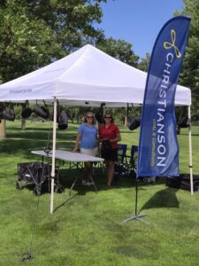 Two individuals stand beneath a tent, with a blue banner with the Christianson CPA logo displayed prominently above them.