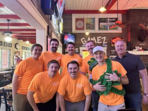 A group of men wearing orange shirts smiles together for a photo, showcasing camaraderie and teamwork.