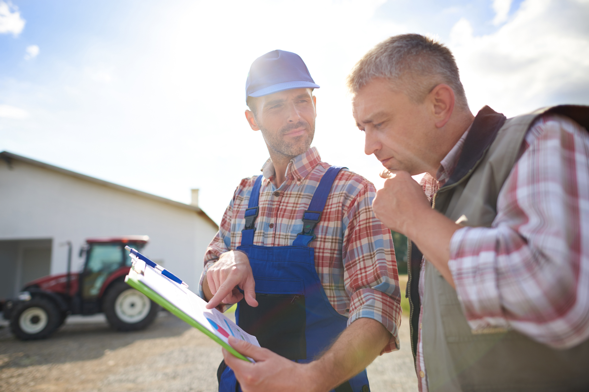 Two men stand in front of a farm, with a tractor visible in the background, showcasing rural life and agriculture.