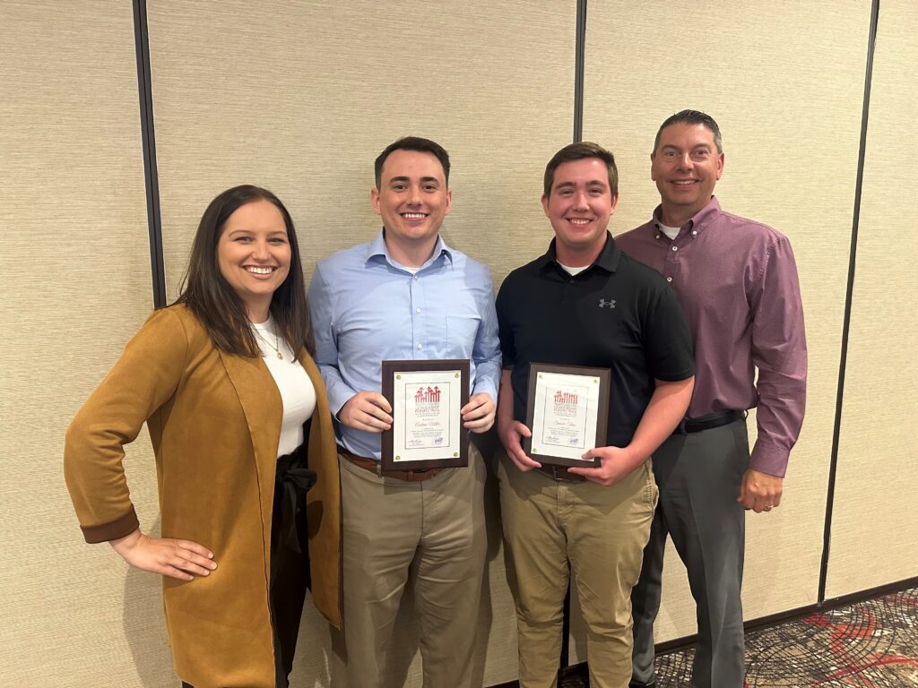 Two men smiling with awards in their hands, surrounded by supportive leadership team