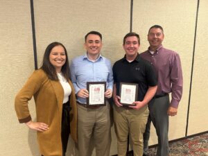 Two men smiling with awards in their hands, surrounded by supportive leadership team