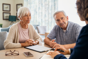The image shows an elderly couple meeting with a professional advisor at a desk. The couple is seated side by side, with the woman on the left and the man on the right. The woman has short white hair, wears a beige cardigan over a pink top, and has her hands clasped in front of her. The man has gray hair, wears a light blue short-sleeved shirt, and is leaning slightly forward, engaged in conversation. They are both attentively listening to the advisor, who is partially visible from behind. On the desk in front of them is a clipboard with documents, a pen, and a smartphone, as well as the woman's glasses. The setting appears to be a well-lit, cozy office or living room with light-colored walls and decor, giving the image a warm and professional atmosphere.