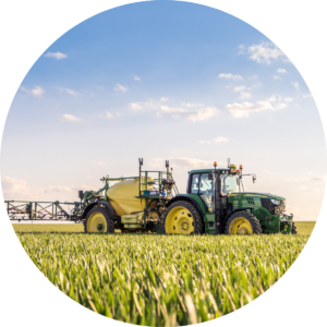 The image is a circular photograph showing a green tractor pulling a large yellow agricultural sprayer through a vibrant, green field of crops. The tractor is positioned to the right, with its large yellow wheels clearly visible. The field stretches out in front, full of tall, healthy crops. Above, the sky is bright blue with scattered white clouds, suggesting a sunny day. The image evokes a sense of modern farming and agricultural productivity.